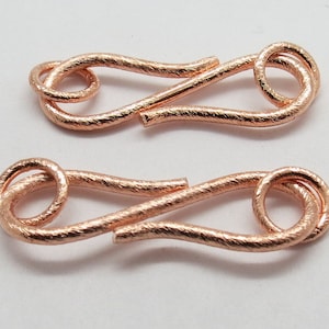 May include: Two rose gold metal clasps with a textured finish. Each clasp has a loop on one end and a hook on the other end.