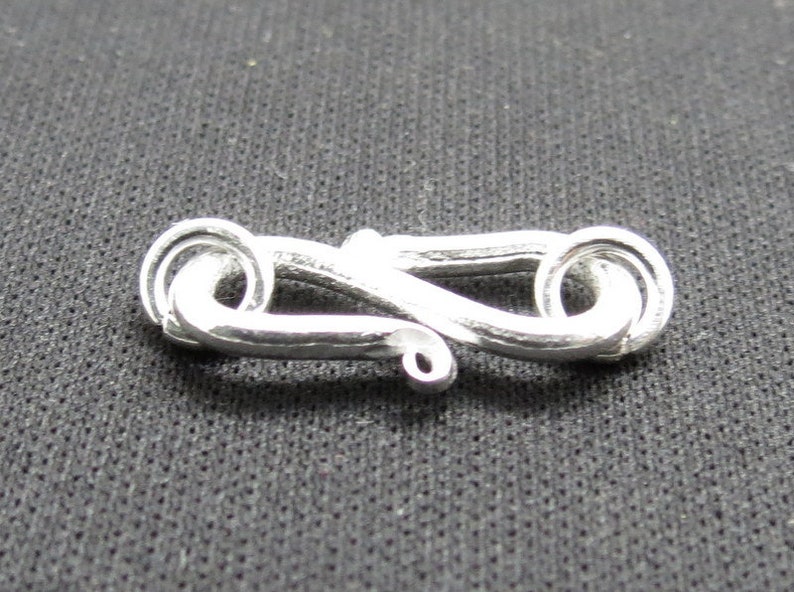 Puede incluir: Cierre de metal plateado con un dise&ntilde;o &uacute;nico e intrincado. El cierre est&aacute; hecho de dos bucles entrelazados.