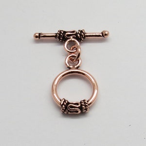 May include: A rose gold toggle clasp with a decorative rope design. The clasp has a round ring and a bar with two small balls on the ends.