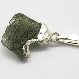 Puede incluir: Un colgante de tektita verde con un engaste y una anilla de plata. La tektita es un vidrio natural formado por el impacto de un meteorito.