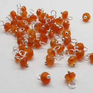 Könnte beinhalten: Ein Haufen orangefarbener Karneol-Perlen mit silbernen Sprungringen. Die Perlen sind facettiert und haben eine polierte Oberfläche.