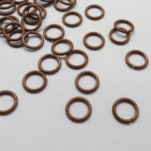 Puede incluir: Un conjunto de pequeños anillos de salto de metal marrón esparcidos sobre una superficie clara. Los anillos son circulares con una pequeña abertura. Estos hallazgos se utilizan a menudo en la fabricación de joyas y artesanías.