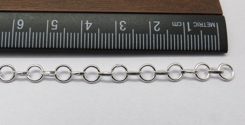 Op de afbeelding: Een close-up van een zilveren ketting met ronde schakels. De ketting wordt gemeten met een liniaal, wat laat zien dat elke schakel ongeveer 5 mm in diameter is.