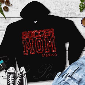 May include: Black hoodie with red glitter text that says "Soccer Mom" and the name "Madison" underneath.