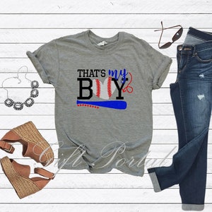 May include: Grey t-shirt with the text "That's my Boy" and a baseball graphic. The shirt is paired with blue jeans, a necklace, and brown wedges.