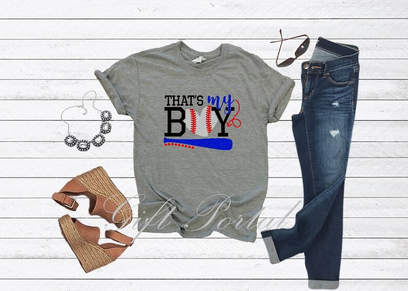 May include: Grey t-shirt with the text "That's my Boy" and a baseball graphic. The shirt is paired with blue jeans, a necklace, and brown wedges.