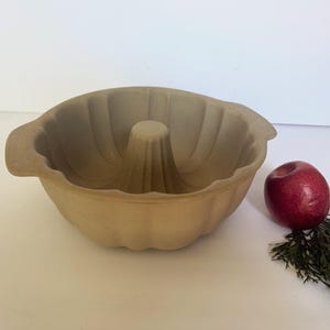 May include: A tan-colored bundt pan with a central cone and fluted sides. A red apple and a sprig of greenery are next to the pan. The pan has two handles and is made of a stoneware material.