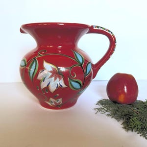 May include: A red ceramic pitcher with a white floral design and a curved handle. The pitcher is decorated with white flowers, green leaves, and blue accents. An apple and greenery are next to the pitcher.