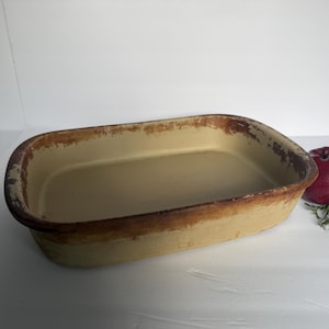 May include: A rectangular baking dish with a cream-colored interior and a distressed, dark brown rim. The exterior features a worn, tan finish with visible signs of use and aging along the edges, showcasing a rustic appearance for kitchen use.