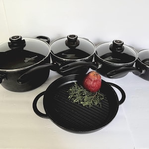 May include: A set of black cookware with glass lids and black handles. The set includes pots and pans of various sizes, along with a grill pan. An apple and herbs are placed in the grill pan.