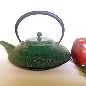 May include: A green cast iron teapot featuring a relief design of elephants walking through tall grass. The teapot has a black arched handle, a matching lid with a small knob, and a short spout. A red apple sits beside the teapot.