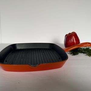 May include: Orange cast iron grill pan with a black nonstick surface and a long handle. The pan is sitting on a white surface.