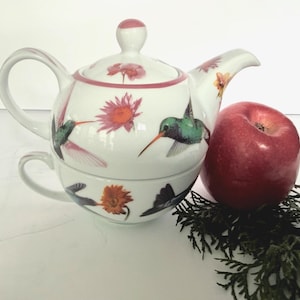 May include: A white ceramic tea-for-one set consisting of a teapot with a lid and a matching teacup, decorated with colorful hummingbirds and orange and pink flowers. A red apple and a small sprig of green evergreen needles sit beside the set.