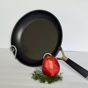 Puede incluir: Una sart&eacute;n antiadherente negra con un mango plateado y un agarre negro. Una manzana roja descansa sobre un lecho de follaje verde junto a la sart&eacute;n. La sart&eacute;n est&aacute; sostenida por un soporte met&aacute;lico decorativo.