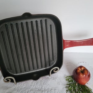 May include: Red cast iron grill pan with a black, ridged cooking surface and a long red handle. The pan is sitting on a white surface.