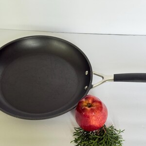 Puede incluir: Una sart&eacute;n antiadherente negra con un mango plateado y un agarre negro. Una manzana roja y ramitas verdes est&aacute;n al lado de la sart&eacute;n. La sart&eacute;n tiene aproximadamente 30 cm de di&aacute;metro.