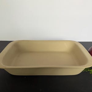 May include: A rectangular, tan-colored baking dish with rounded corners. A red apple and green sprigs are to the right of the dish. The dish is on a black surface.