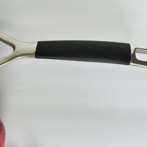 Puede incluir: Primer plano del mango de una sart&eacute;n negra con un soporte de metal plateado. El mango tiene un agarre de goma negra y un agujero en el extremo para colgar. Una manzana roja es visible en la esquina inferior izquierda.