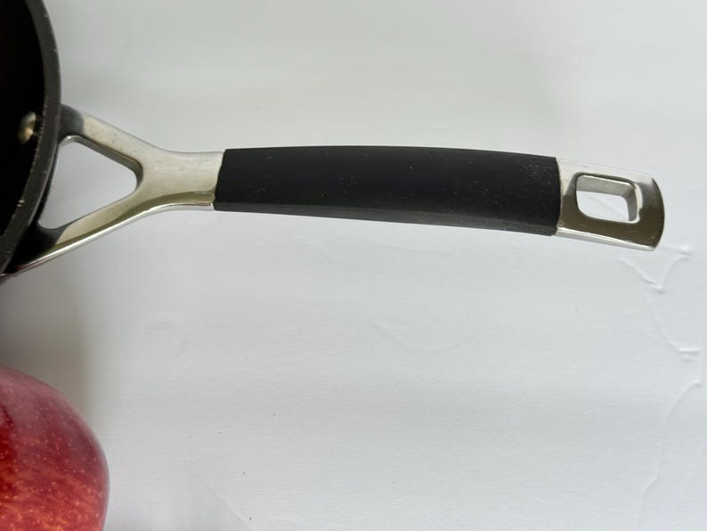 Puede incluir: Primer plano del mango de una sart&eacute;n negra con un soporte de metal plateado. El mango tiene un agarre de goma negra y un agujero en el extremo para colgar. Una manzana roja es visible en la esquina inferior izquierda.