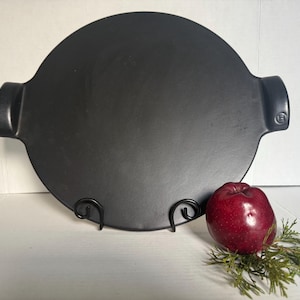 May include: A round, matte black ceramic serving tray with two integrated side handles. The tray features a small embossed logo on the right handle. It is displayed upright on a black metal stand, accompanied by a single red apple and greenery.
