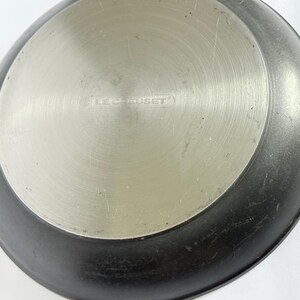 Puede incluir: Primer plano de la base de una sart&eacute;n Le Creuset negra y plateada. El centro es plateado con el nombre de la marca grabado en el metal. El borde exterior de la sart&eacute;n es negro.