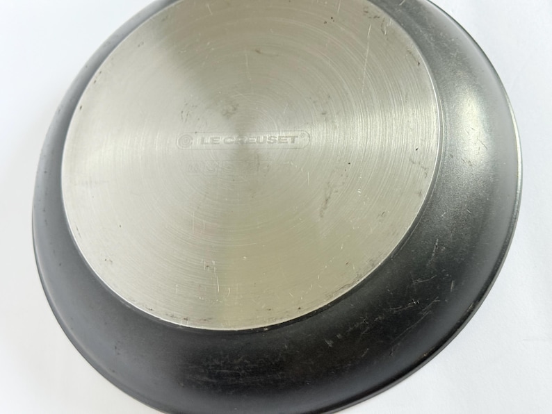 Puede incluir: Primer plano de la base de una sart&eacute;n Le Creuset negra y plateada. El centro es plateado con el nombre de la marca grabado en el metal. El borde exterior de la sart&eacute;n es negro.
