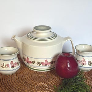 May include: A cream-colored ceramic teapot with a matching lid and two mugs, all featuring a horizontal band of pink floral motifs and thin blue and green stripes. The teapot has a wide base, a curved handle, and a short spout, while the mugs are rounded.