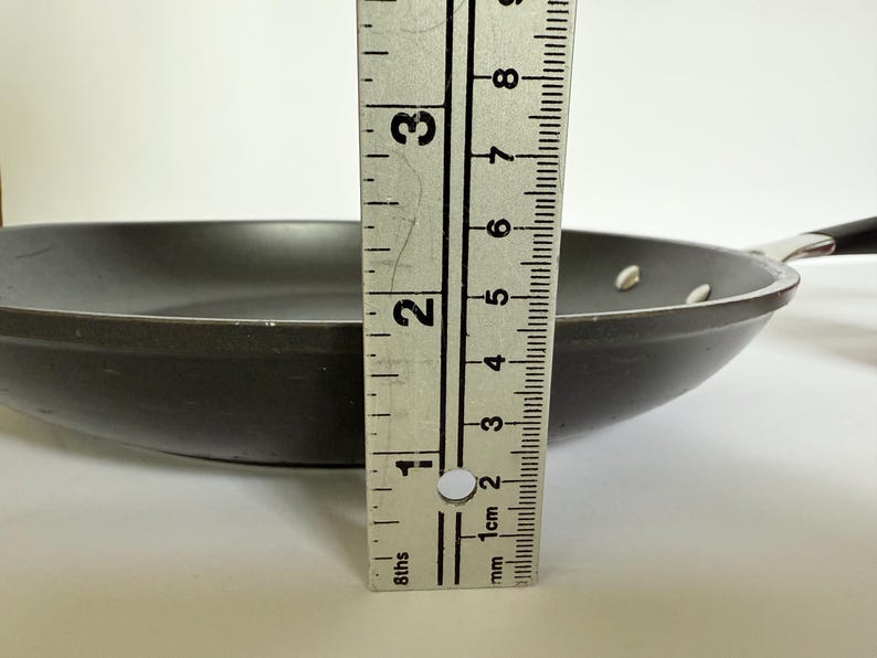 Puede incluir: Una sart&eacute;n gris oscuro con un mango negro. La sart&eacute;n tiene aproximadamente 5 cm de profundidad. Se coloca una regla met&aacute;lica junto a la sart&eacute;n para mostrar la profundidad.