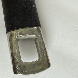 Puede incluir: Primer plano de un mango negro con un extremo de metal plateado. El extremo met&aacute;lico tiene una abertura rectangular. El mango parece ser parte de un utensilio o herramienta de cocina.