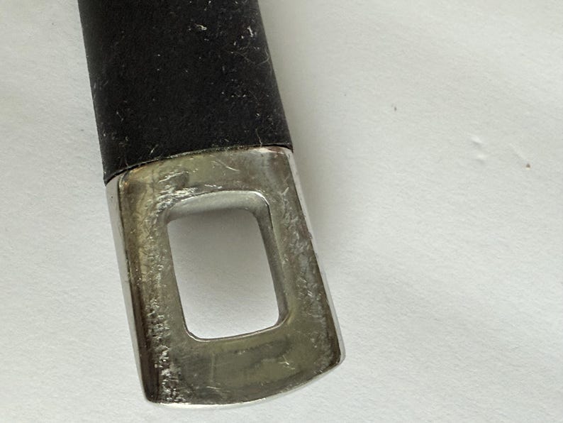 Puede incluir: Primer plano de un mango negro con un extremo de metal plateado. El extremo met&aacute;lico tiene una abertura rectangular. El mango parece ser parte de un utensilio o herramienta de cocina.