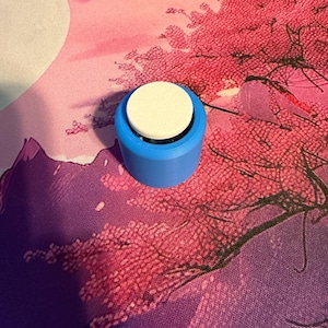 Könnte beinhalten: Ein zylindrisches blaues Kunststoffobjekt mit einer flachen, kreisförmigen weißen Oberseite. Der Gegenstand verfügt über eine glatte, einfarbig blaue Basis und eine leicht vertiefte weiße Scheibe auf der Oberseite, was ein schlichtes Design ergibt.