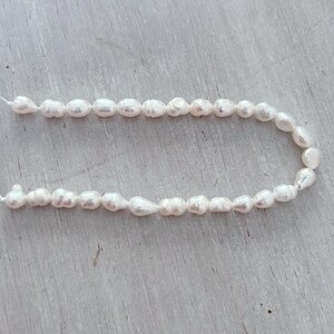 May include: A string of white, irregularly shaped freshwater pearls. The pearls are strung on a thin, white cord, creating a delicate and elegant appearance. The pearls have a natural lustre and varying sizes, suggesting a handmade or organic origin.