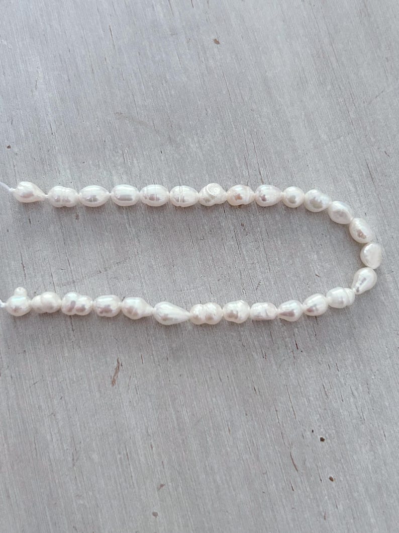 May include: A string of white, irregularly shaped freshwater pearls. The pearls are strung on a thin, white cord, creating a delicate and elegant appearance. The pearls have a natural lustre and varying sizes, suggesting a handmade or organic origin.