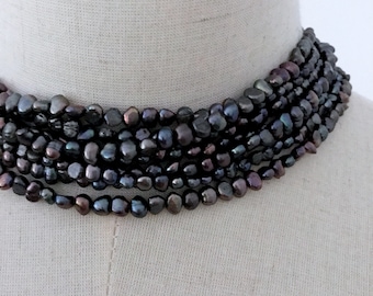 May include: A multi-strand necklace of dark, iridescent pearls. The pearls are irregularly shaped, appearing a deep, lustrous black with hints of purple and green. The necklace is displayed on a mannequin.