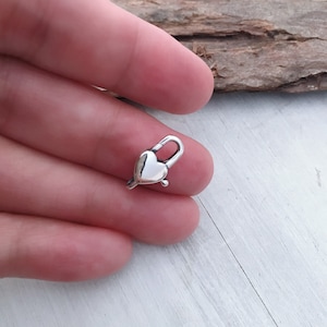 Könnte beinhalten: Silberner herzförmiger Verschluss für die Schmuckherstellung. Der Verschluss ist klein und zart, perfekt, um Halsketten, Armbändern oder Fußkettchen einen Hauch von Eleganz zu verleihen.