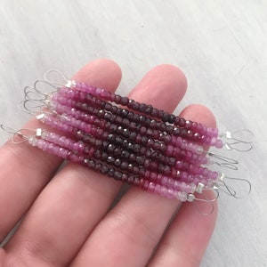 May include: A strand of pink and red gemstone beads, with a gradient of color from light pink to dark red. The beads are strung on a silver wire and have a loop at each end.
