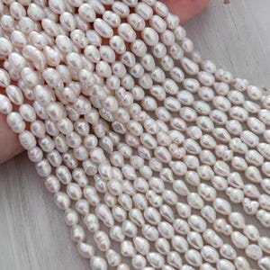 May include: Close-up of multiple strands of creamy white, oval pearls. The pearls are strung together, creating a flowing, textured appearance. The pearls have a natural lustre and varying sizes, suggesting a handmade or natural origin. The background is a light colour.