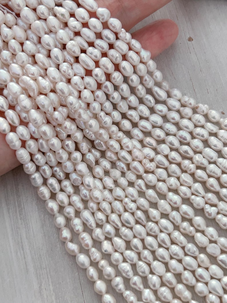 May include: Close-up of multiple strands of creamy white, oval pearls. The pearls are strung together, creating a flowing, textured appearance. The pearls have a natural lustre and varying sizes, suggesting a handmade or natural origin. The background is a light colour.
