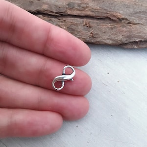 Puede incluir: Un pequeño cierre plateado en forma de infinito, probablemente para la fabricación de joyas. El cierre tiene forma de símbolo de infinito, con un diseño curvo y una pequeña palanca para abrir y cerrar. El cierre se sostiene en una mano.