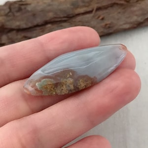 May include: A polished, elongated gemstone with a pale blue and brown pattern. The stone is held in a hand, showcasing its smooth, translucent surface and natural color variations. The gemstone's shape is reminiscent of a leaf or a teardrop.