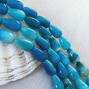 May include: A strand of teal blue agate gemstone beads in a teardrop shape. The beads have white and light blue striations.