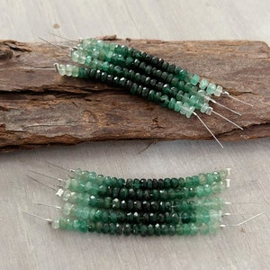 May include: A strand of green gemstone beads with a gradient of color from light to dark green. The beads are faceted and have a square shape. The beads are strung on a thin wire.