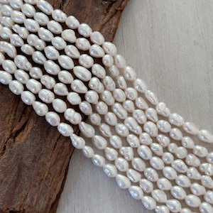 May include: Close-up of multiple strands of creamy white freshwater pearls. The pearls are oval-shaped with a smooth, glossy finish. The strands are arranged in parallel lines, with a piece of dark brown wood in the background.