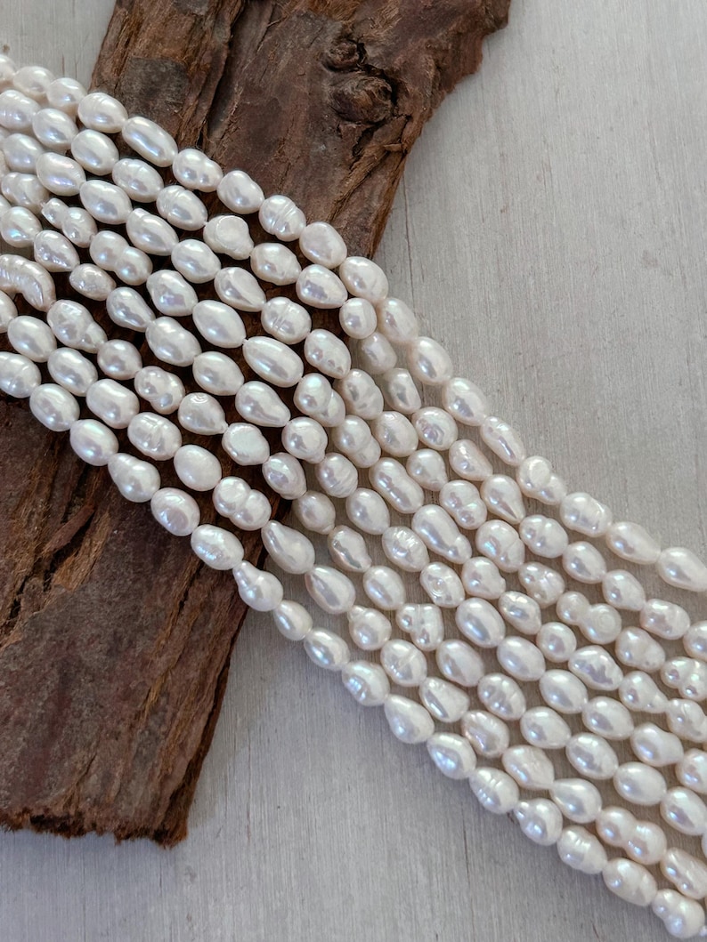 May include: Close-up of multiple strands of creamy white freshwater pearls. The pearls are oval-shaped with a smooth, glossy finish. The strands are arranged in parallel lines, with a piece of dark brown wood in the background.