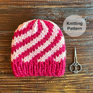 May include: A pink and white striped knitted hat on a wooden surface. The hat has a diagonal stripe pattern. Knitting Pattern is written on a white circle.