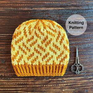Puede incluir: Un gorro de punto con un patrón geométrico en amarillo y blanco. El gorro está sobre una superficie de madera con unas tijeras a su lado. El texto "Knitting Pattern" está escrito en un círculo blanco.