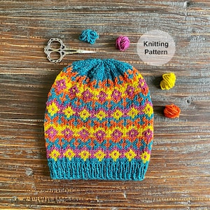 May include: A teal blue knitted hat with a colourful geometric pattern in yellow, orange, pink, and purple. The hat is on a wooden surface with yarn balls in various colours.