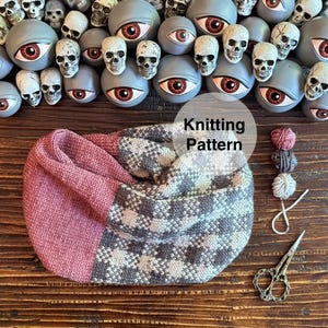 May include: A knitted cowl in shades of pink, grey, and white, with a geometric pattern. The text "Knitting Pattern" is visible. Small balls of yarn and scissors are next to the cowl. Decorative skulls and eye-shaped objects are in the background.