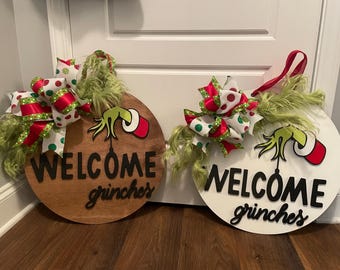May include: Two round wooden door signs with the text "WELCOME grinches". One sign is brown, the other is white. Each has a Grinch hand graphic and a decorative bow with red, green, and white accents.