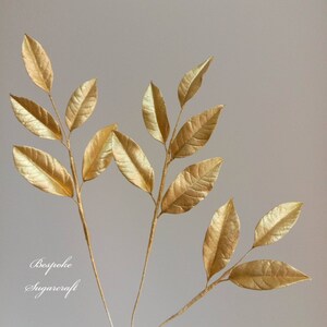 Three gold sugar leaf sprigs with multiple leaves on each stem. The leaves are a metallic gold colour and have a slightly textured surface. The stems are thin and brown. The image has the text "Bespoke Sugarcraft" at the bottom.
